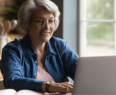 Focused happy elderly 60s grey-haired woman in glasses look at laptop screen work online at home. Smart modern mature Caucasian female busy using computer, browse internet or pay bills on web.